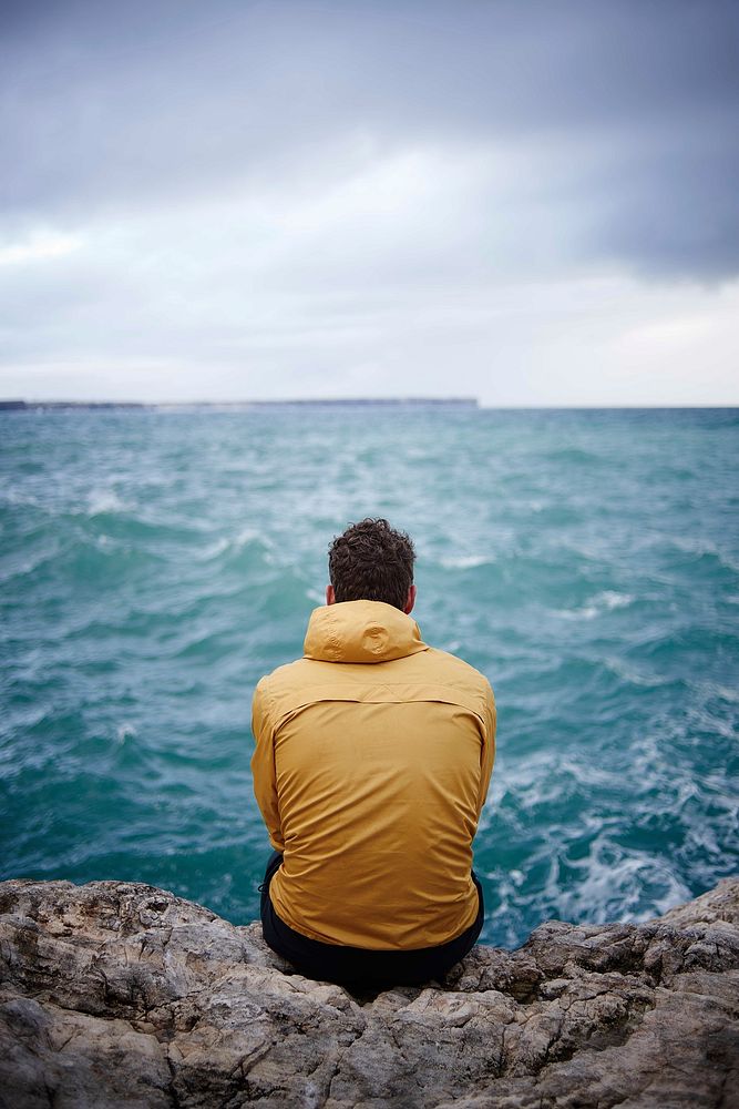 Man sitting on cliff facing ocean, free public domain CC0 photo.