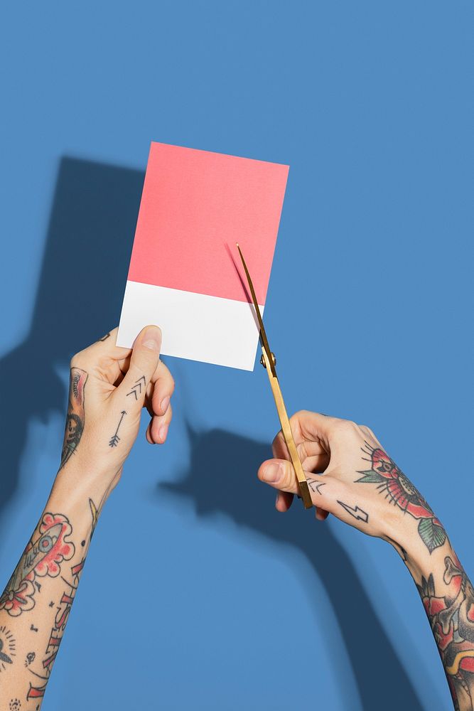 Tattooed hands cutting a red paper with a blue wall