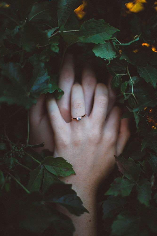 A ring is highlighted on a young woman's hand clasped over her face behind vines. Original public domain image from…