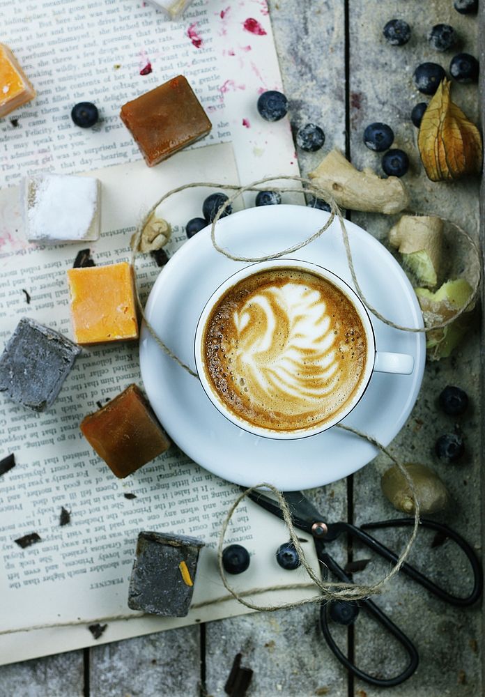 An overhead shot of a cup of coffee with latte art surrounded by blueberries and various trinkets. Original public domain…
