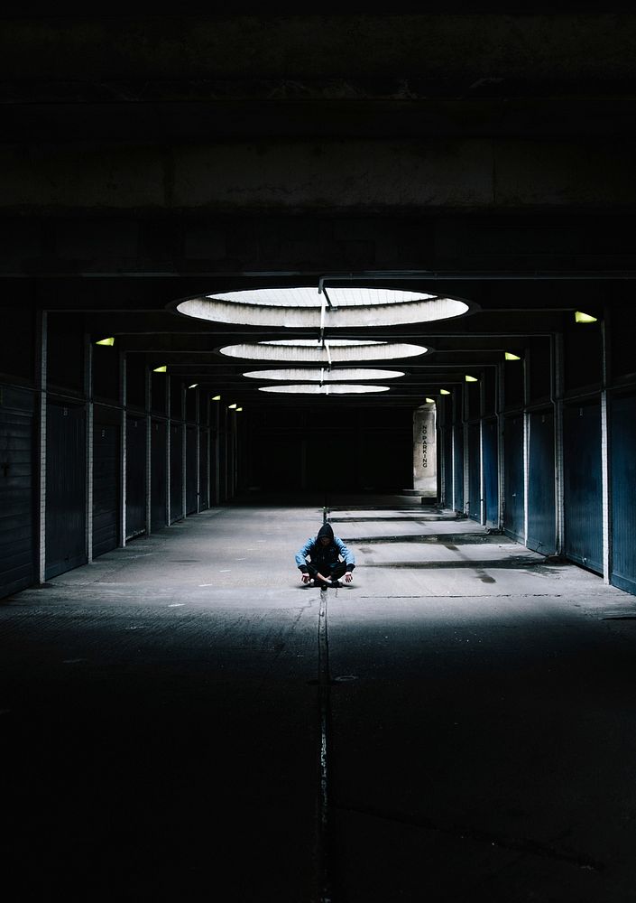 A man in a hoodie sits on the floor of a deserted parking lot in London.. Original public domain image from Wikimedia Commons