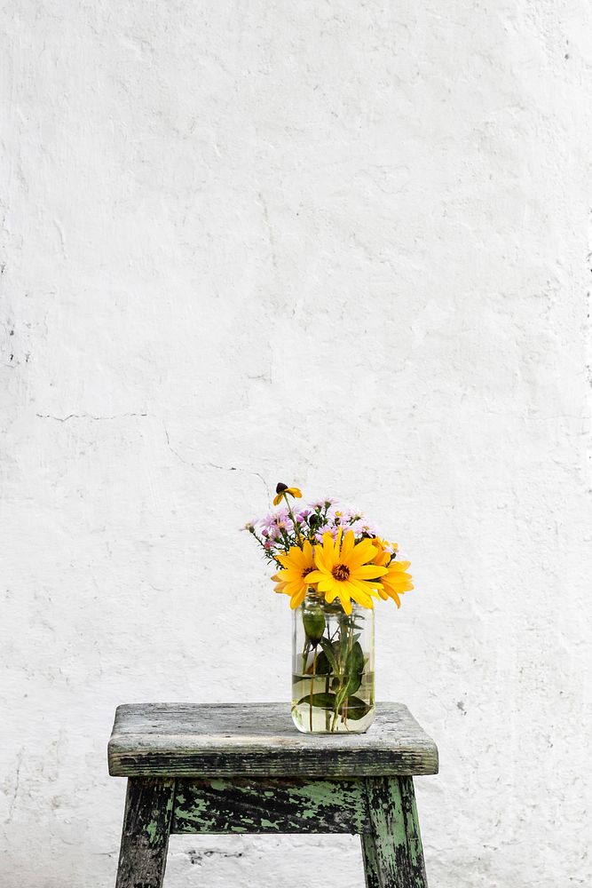 Glass vase with large yellow and small pink flowers on a worn-out stool against an old white wall. Original public domain…