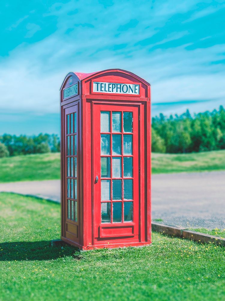 A red telephone booth sits on green grass against a blue sky. Original public domain image from Wikimedia Commons