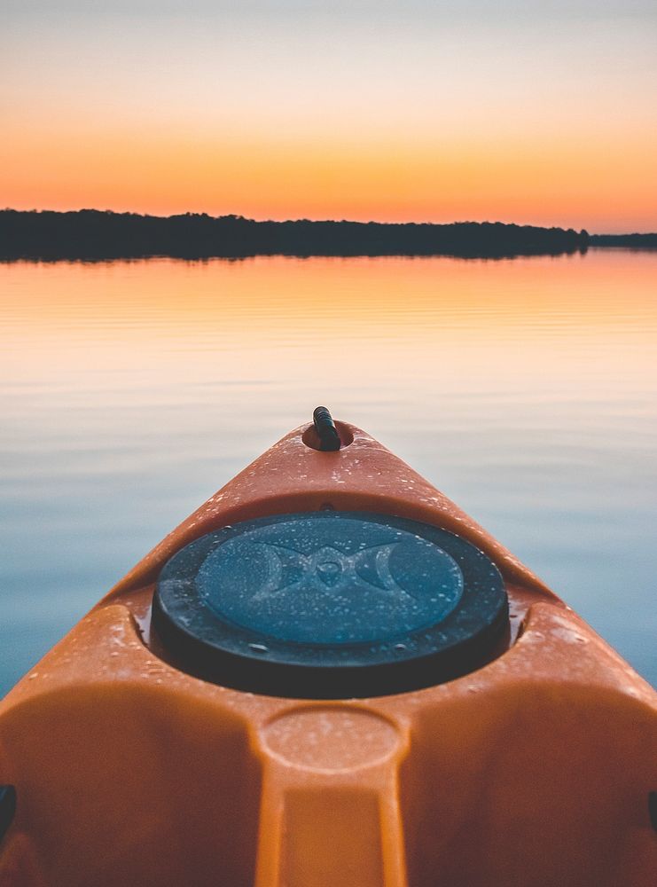 Front point of an orange and black kayak on calm water, trees in the distance, at sunset. Original public domain image from…