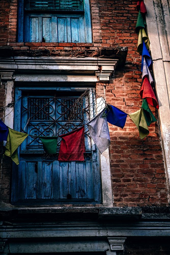 A brick building with blue window shutters and colored flags around it in Swayambhu Stupa. Original public domain image from…