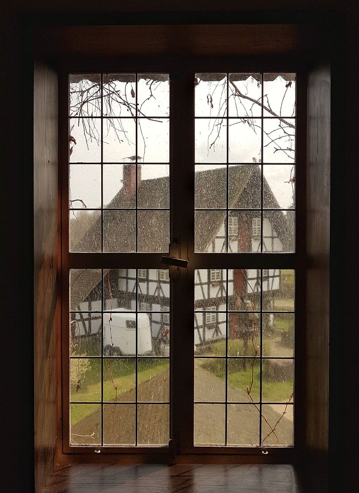 House with a wooden frame and thatched roof seen through a window of a different house. Original public domain image from…