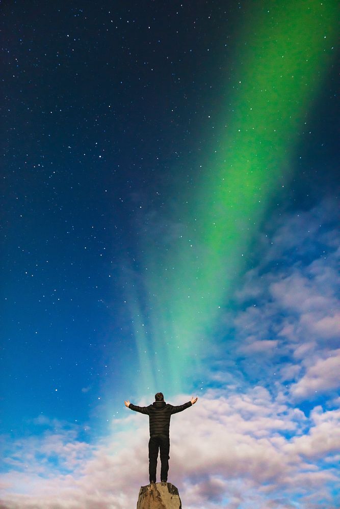 A man raising his arms on top of a rock formation while staring out at a Northern Lights beam in Iceland as it appears to…