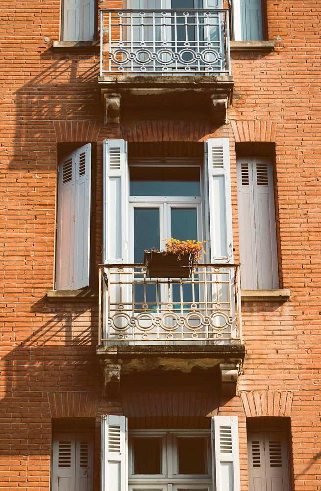 A brick building with large windows that have white shutters and balconies with flower planters on them. Original public…