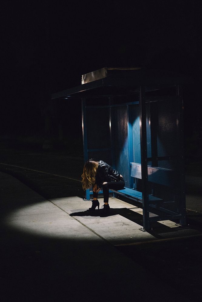 A woman with her head down being illuminated with the only lights of the bus stop.. Original public domain image from…