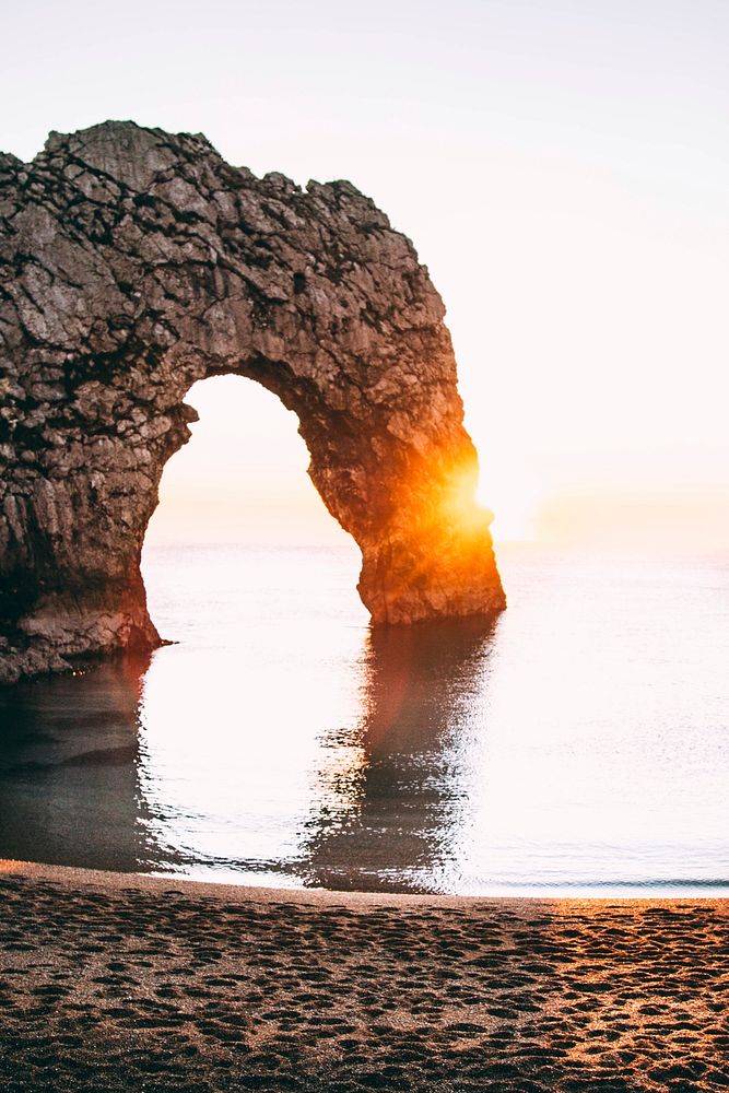 A natural rock arch is illuminated with the rays of a golden sunset over the water near the shoreline of Durdle Door.…
