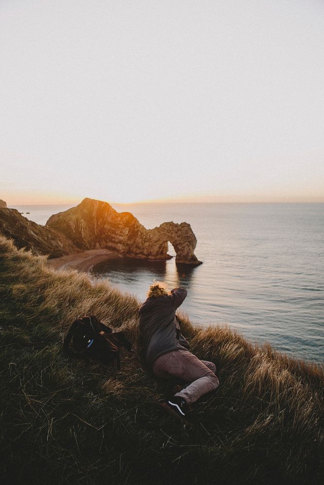 Photographer lying on the grass capturing the Durdle door arch on the cove during golden hour. Original public domain image…