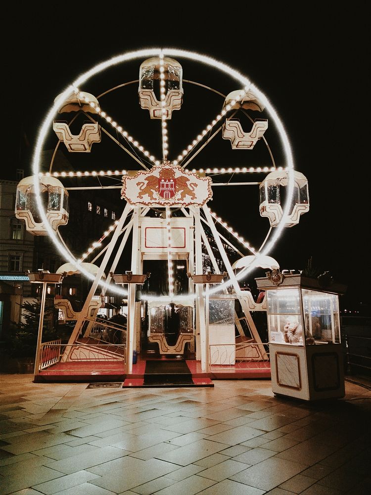 Small ferris wheel lit by bulbs at night with a person waiting in a booth. Original public domain image from Wikimedia…