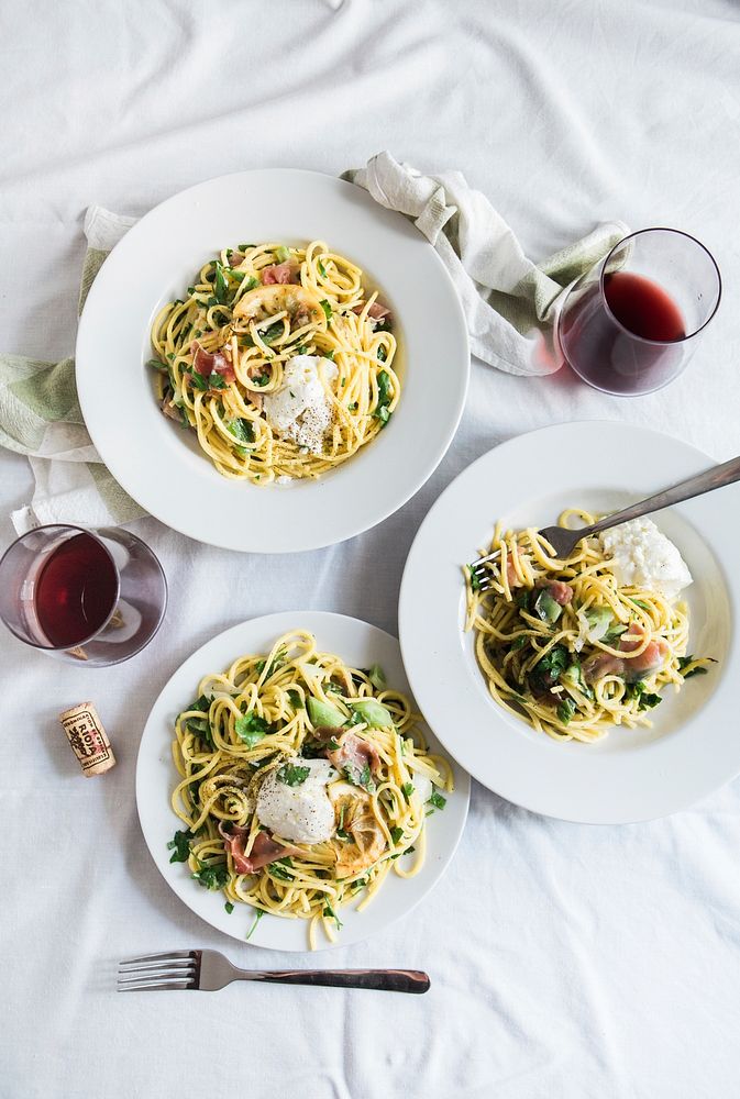 An overhead shot of two glasses of red wine next to three plates of pasta with pancetta on a white tablecloth. Original…