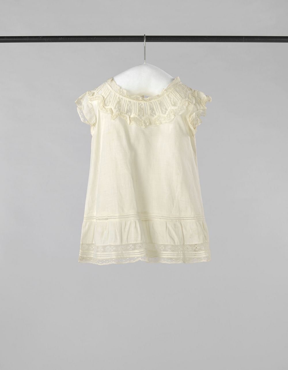 Kinderschort van dun wit katoen, langs bovenlijf, mouwkap, hals en zoom versierd met kant en met een tussenzetsel van kant…