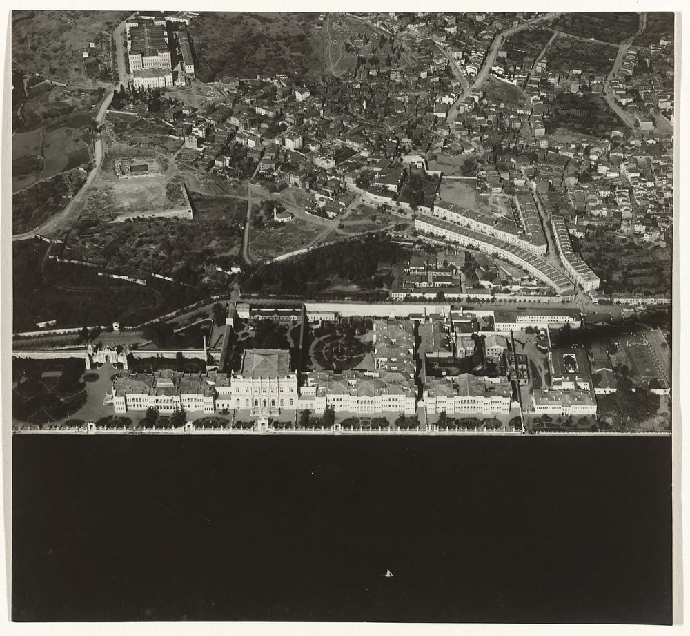 Luchtfoto in zwart-wit van een stad of dorp gelegen aan het water, met een lange U-vormige rij gebouwen (1930 - 1975) by…