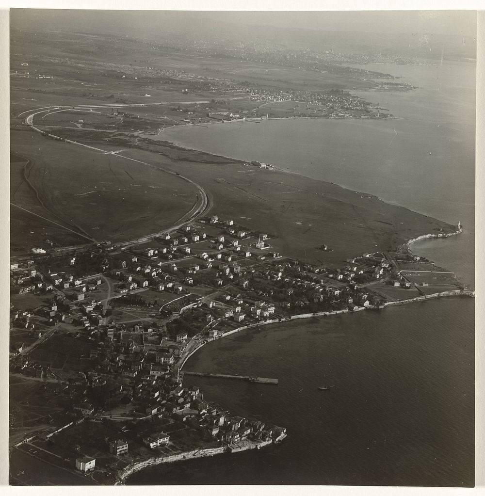 Luchtfoto in zwart-wit van een stad of dorp, gelegen aan het water, met weilanden op de achtergrond, gelegen aan een baai…