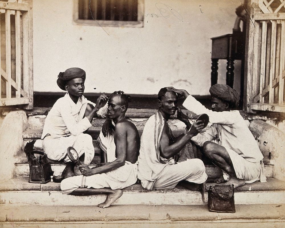 Two Indian barbers wearing turbans shaving the heads of two Indian men who are sitting back to back on some stone steps.…