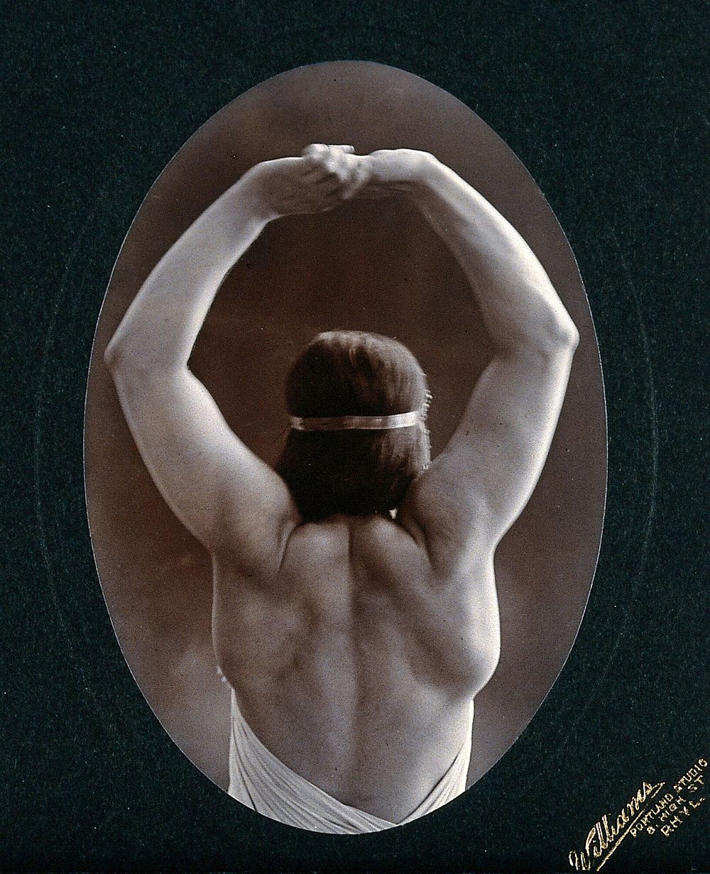 A young woman posing semi-naked, back view, with her arms in the air, in a photographic studio. Photograph by Portland…