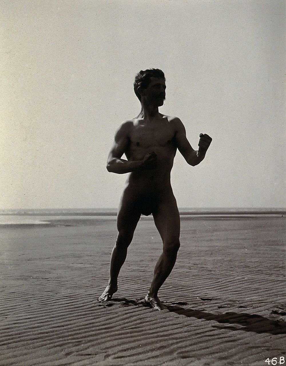 A man posing naked in a boxing stance with his fists raised, on a deserted beach.