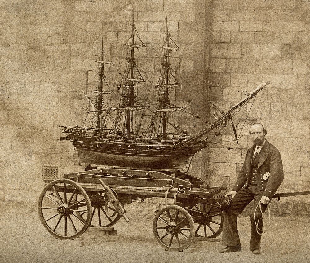 James Pullen, sitting on the end of a wooden trolley upon which rests a huge model ship made by him. Process print.