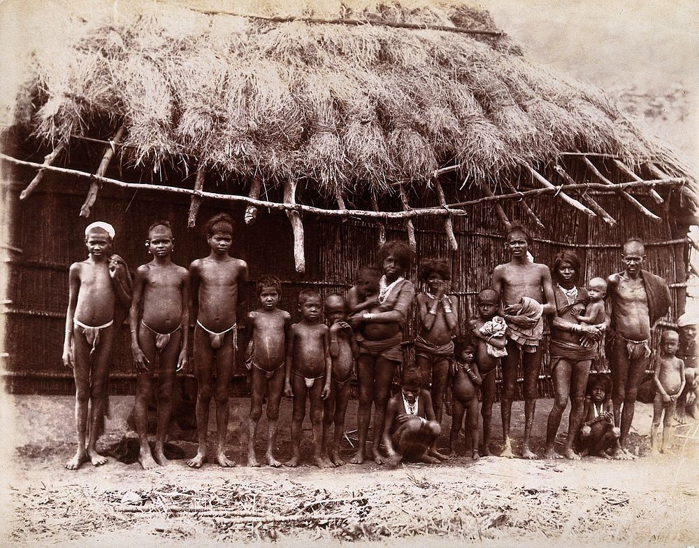 A group of adults and children standing in a line outside a bamboo hut with a thatched roof: Bombay at the time of the…