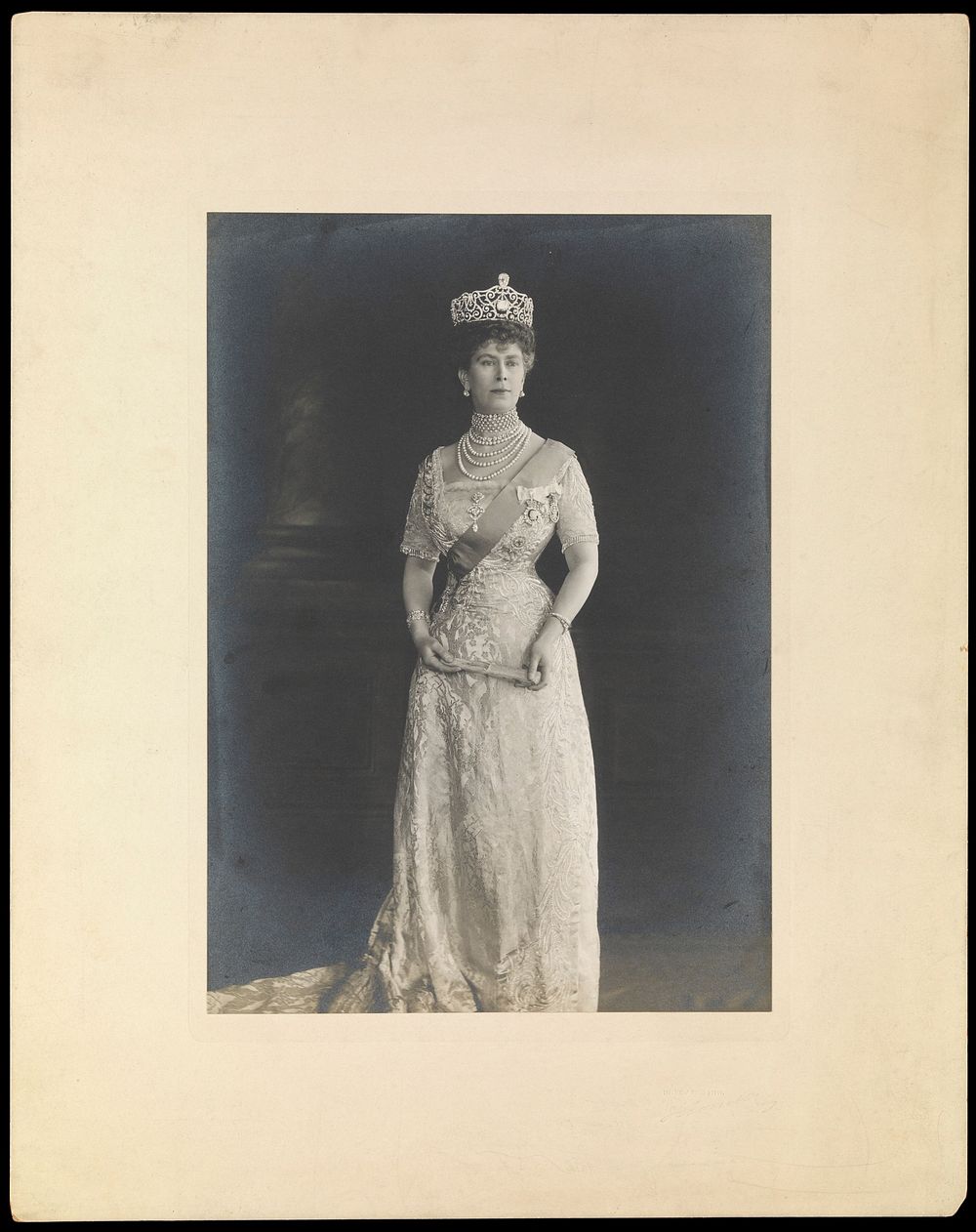Queen Mary (May of Teck) standing, wearing a tiara and holding a folded fan. Photograph by John Thomson, ca. 1910.