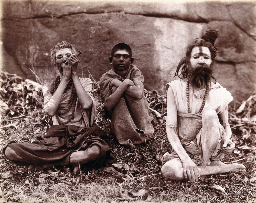 Two men and a boy sitting cross-legged on the ground surrounded by leaves; their faces are painted white and one of them…
