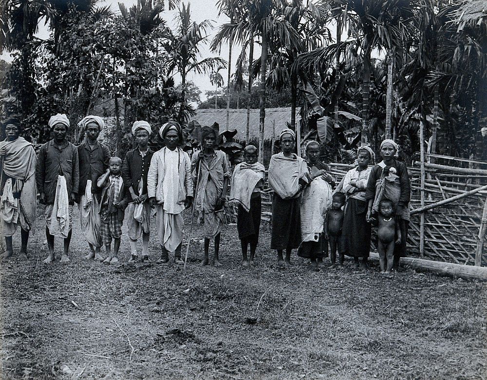 India: Aituni people standing in a row, with palm trees and a grass-roofed house behind them. Photograph, 1900/1920 .