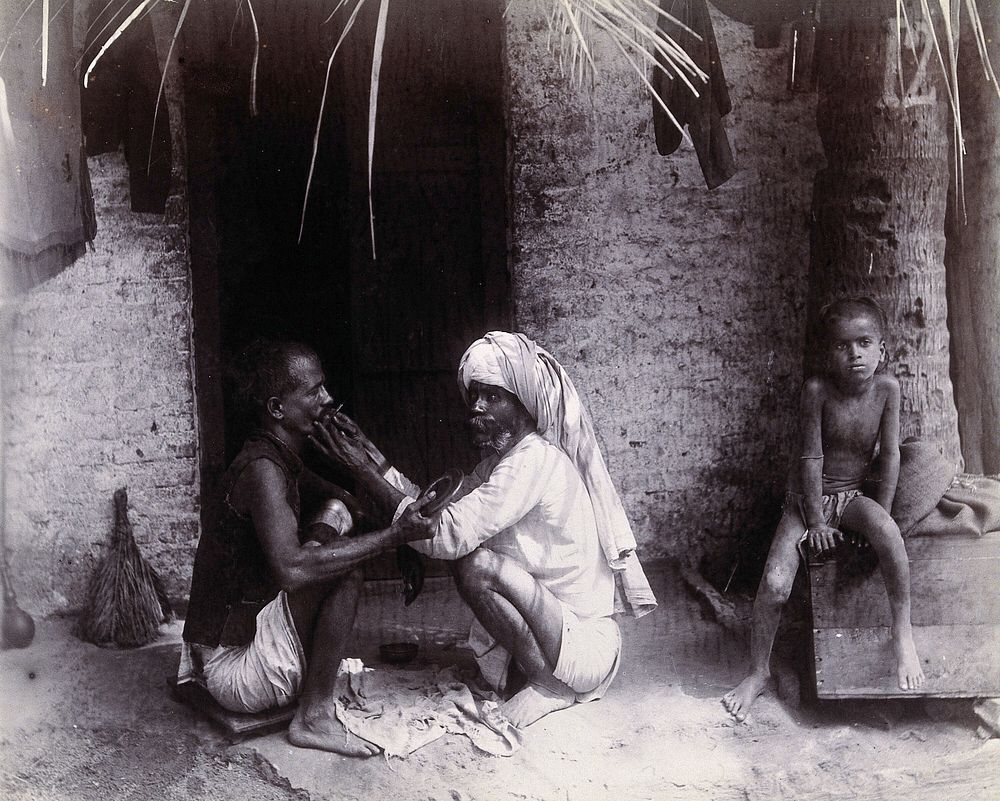 Two Indian men crouching; one is cutting the other's hair, while a girl sits on a wooden box below a tree with the number 12…