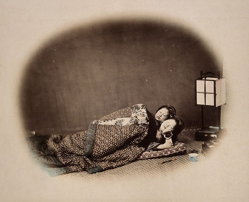 Two Japanese women asleep under a quilt. Coloured photograph by Felice Beato, ca. 1870.