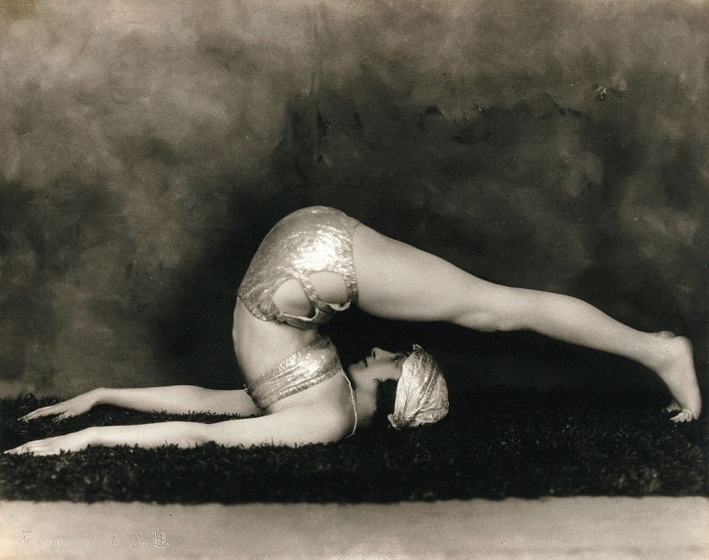 Marguerite Agniel posing with her back arched and legs stretched out over her head, wearing a two-piece costume and matching…