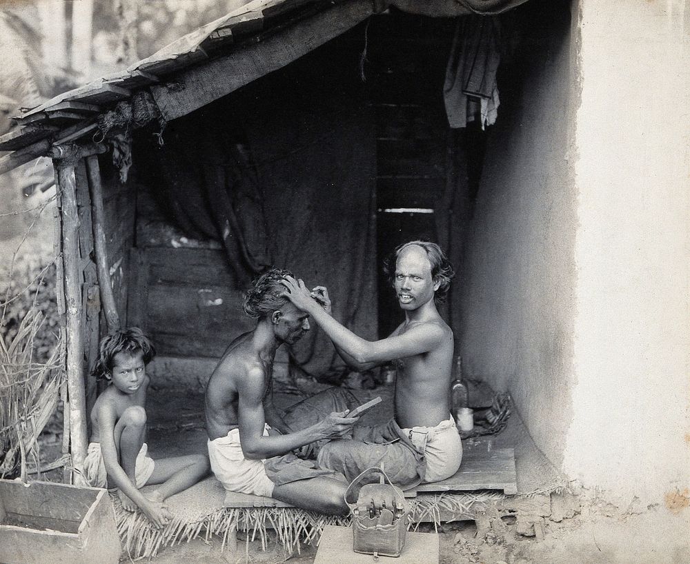 Two men sitting cross-legged; one is a barber shaving the front of the other's head. Photograph.