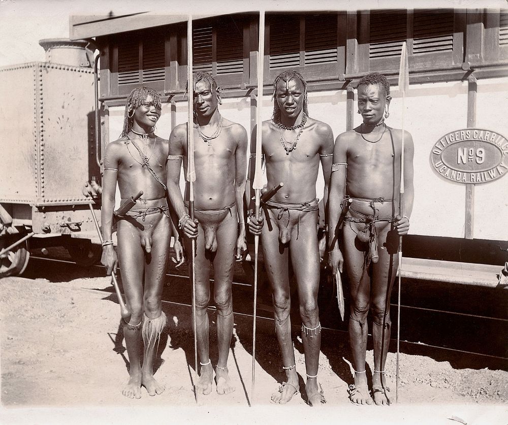 Four young Maasai men, carrying spears, standing beside a train. Photograph, ca.1900.