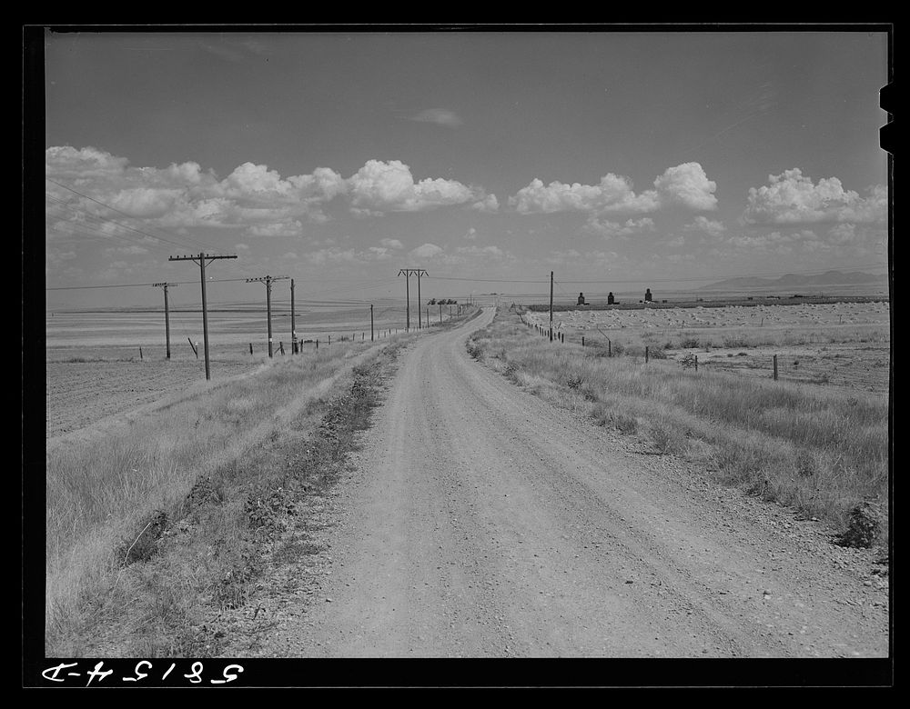 Road to Portage, Montana, with stacks of wheat and grain storage elevators in distance on right. Sourced from the Library of…