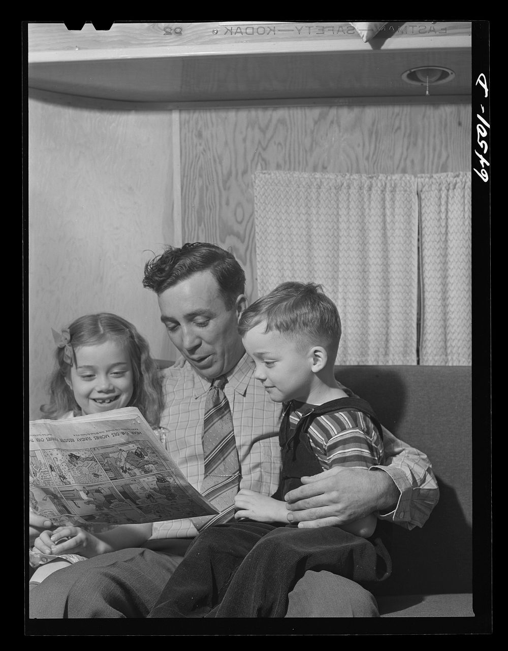 Burlington, Iowa. Cecil Patrick reading the funnies to his kids. FSA (Farm Security Administration) trailer camp for workers…