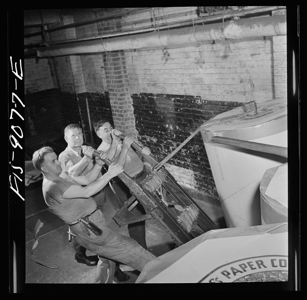 New York, New York. Reel room of the New York Times newspaper. Transporting rolls of paper to presses. Each roll weighs…