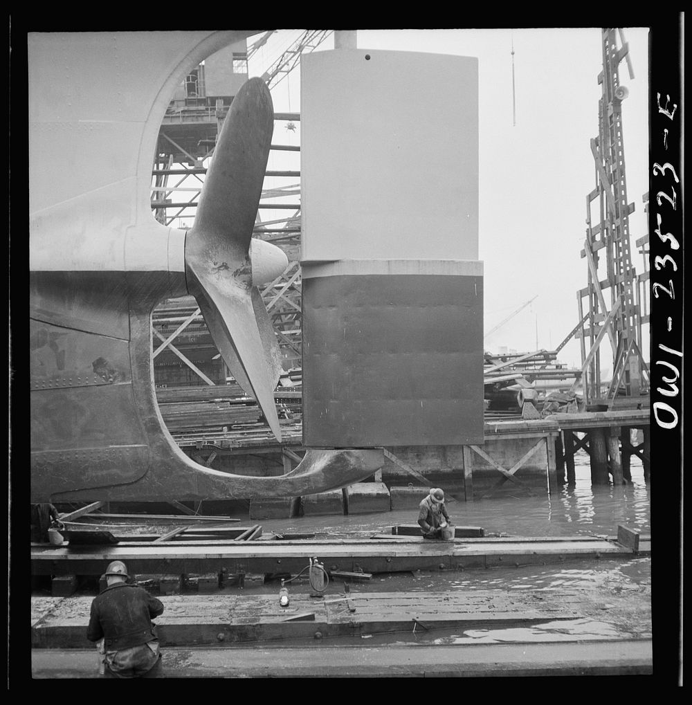 Bethlehem-Fairfield shipyards, Baltimore, Maryland. Propeller and rudder just before a launching. Below, workmen are…