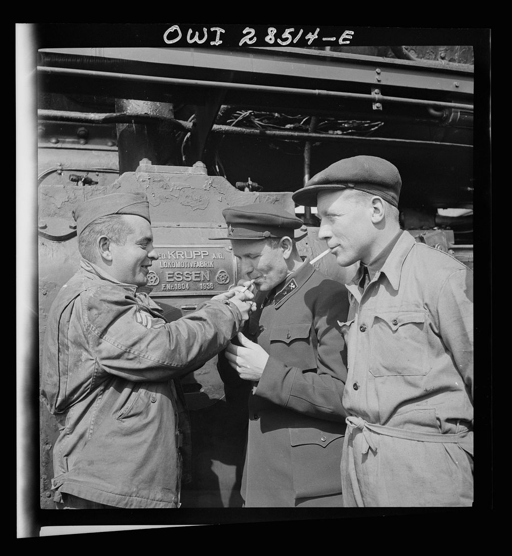 An American Sergeant railroader lighting the cigarette of two Russian railroad engineers as they stand before a German-made…