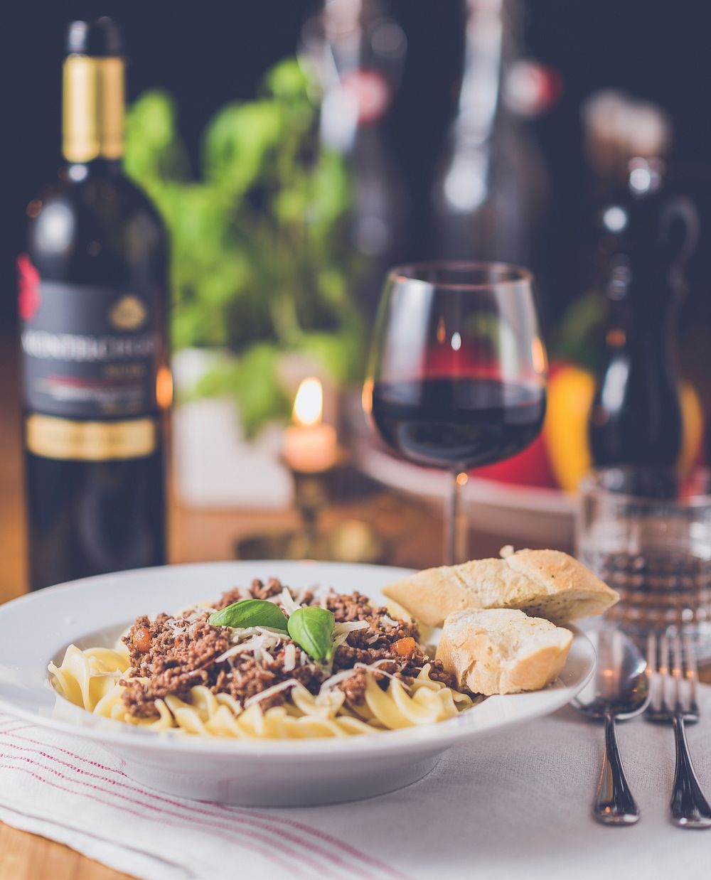 A plate with a pasta dish next to a wine bottle and a glass of red wine on a set table. Original public domain image from…