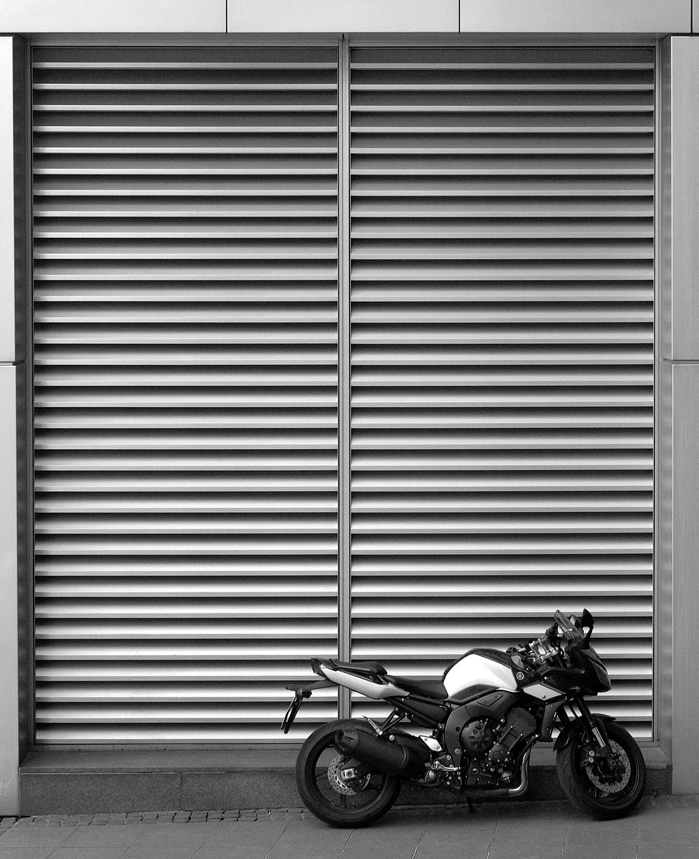 Black and white shot of motorbike parked in front of storefront shutters. Original public domain image from Wikimedia Commons