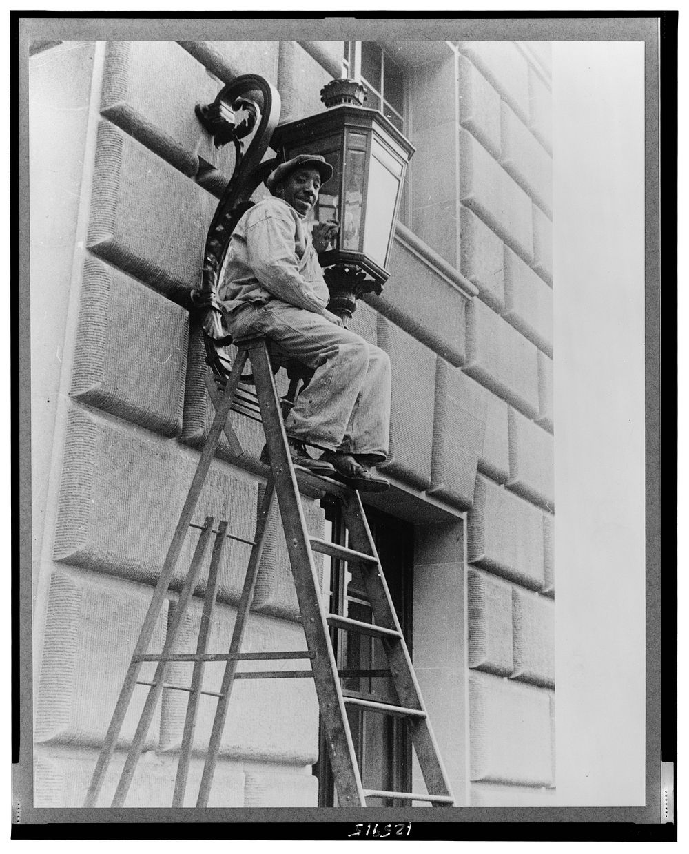 Washington, D.C. A government laborer on a stepladder cleaning a lamp above the entrance of the Commerce(?) building.…