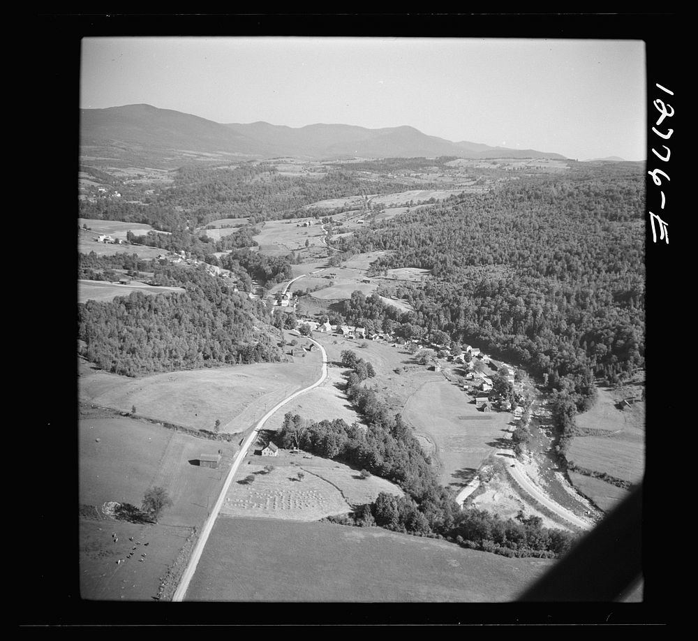 Air view. Dairy farm with hay stacks and cows in foreground. Lincoln, Vermont in distance. Lincoln, Vermont. Sourced from…