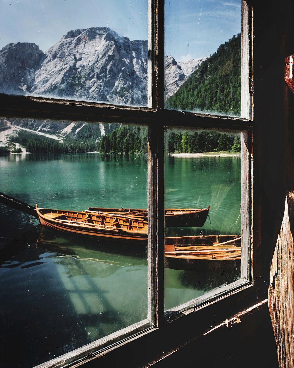 A window view looking at a pair of boats floating in the river with a mix of green and snow covered mountains.. Original…