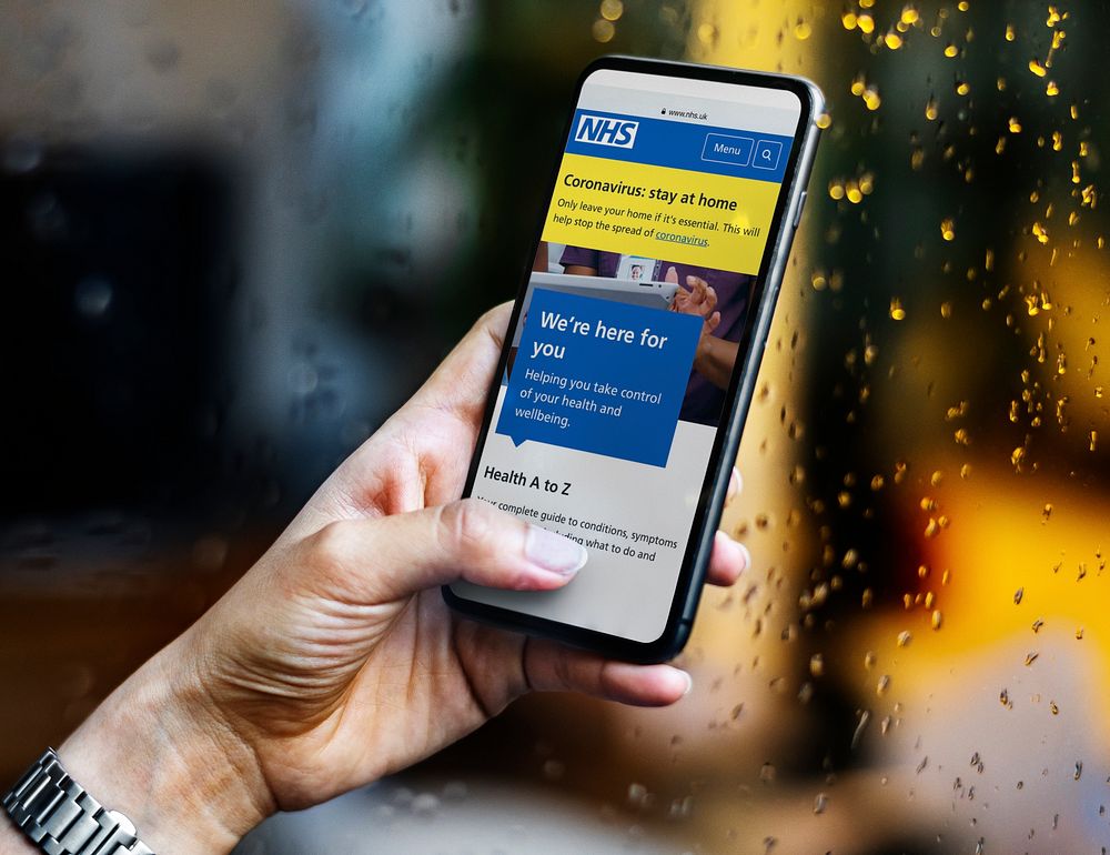 Woman reading coronavirus updates from a mobile phone mockup with editorial graphic from https://www.nhs.uk accessed on…