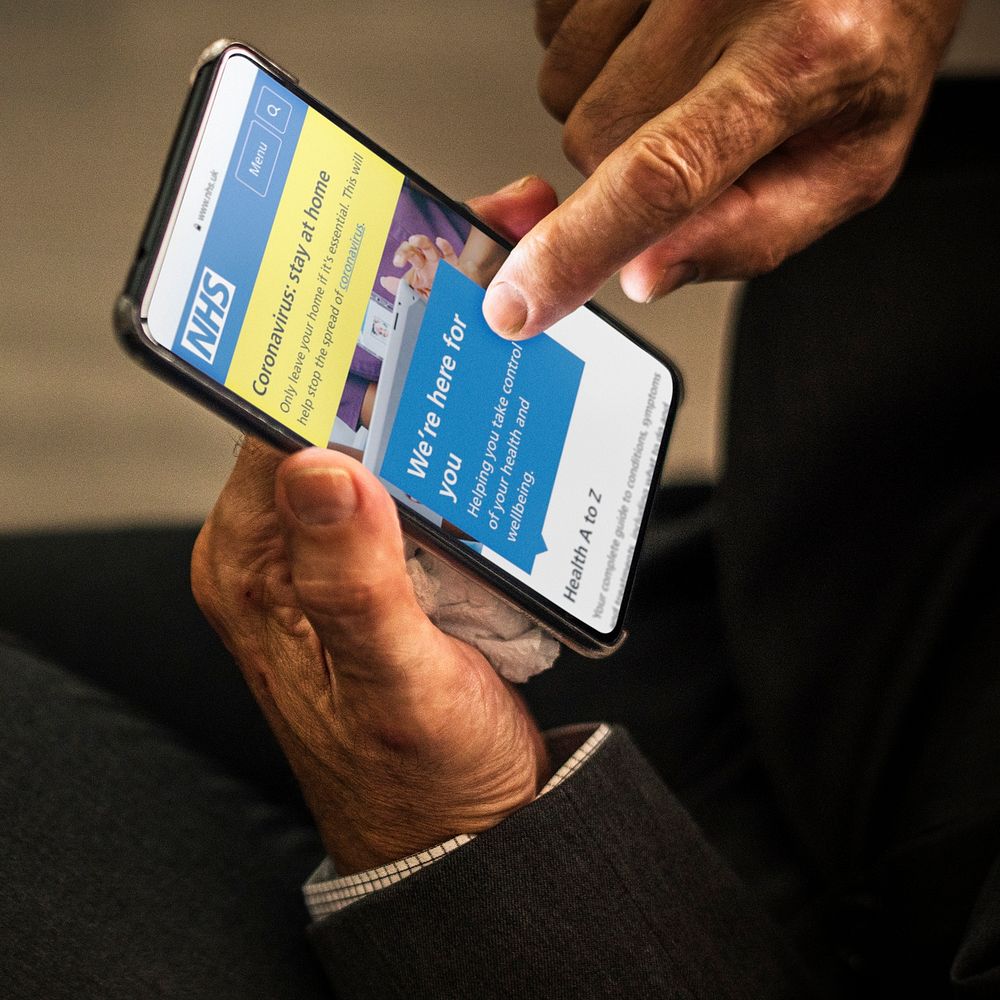 Businessman reading coronavirus updates from a mobile phone mockup with editorial graphic from https://www.nhs.uk accessed…