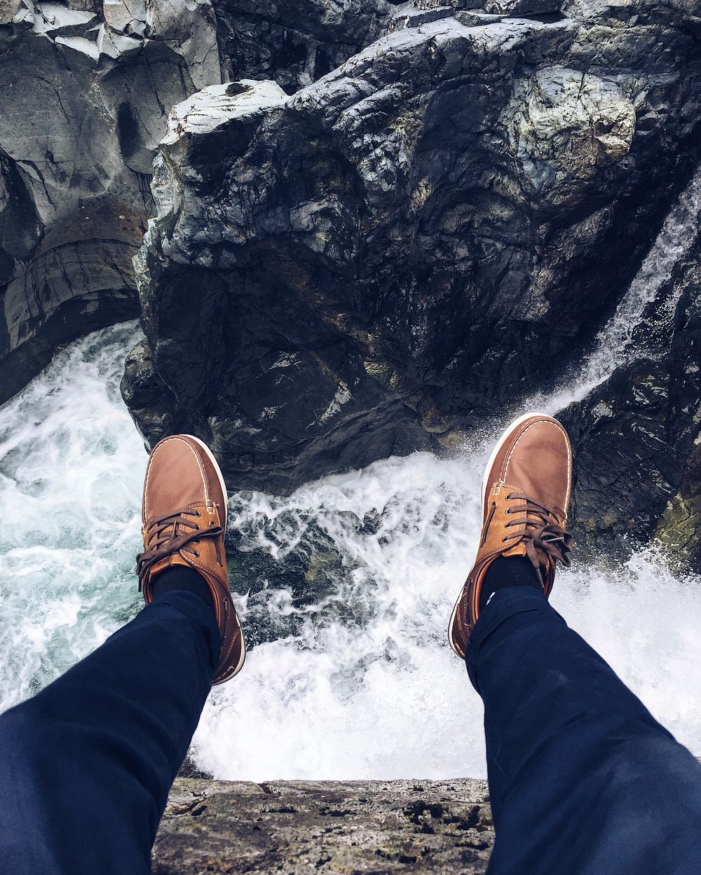 A person's feet in leather shoes dangling off a rock ledge over a fast-flowing creek. Original public domain image from…