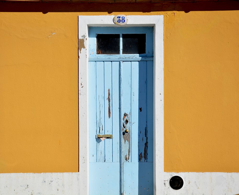 Old wooden door with chipped blue paint with a brass doorknob and the number 38 at the top surrounded by mustard yellow…