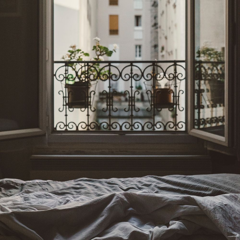 An open window at the foot of an bed with messy sheets looks at flowers on the sill and other buildings across the way.…