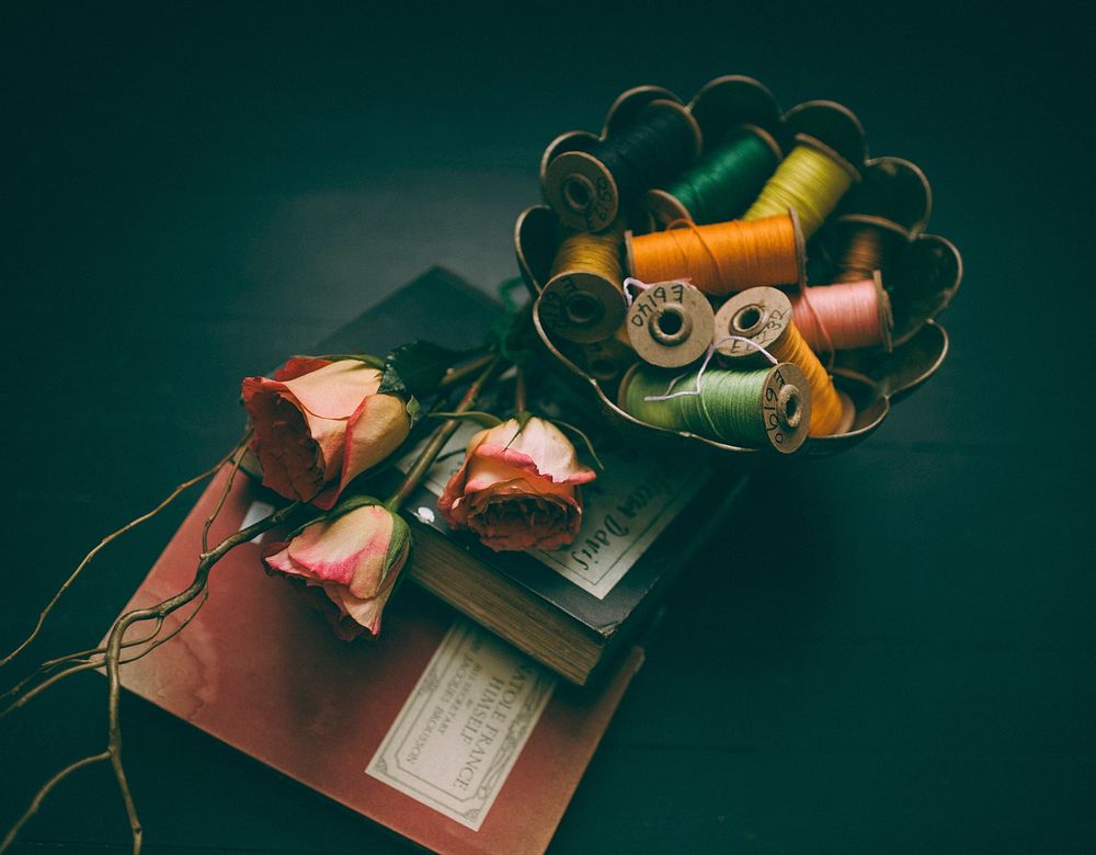 Colorful spools of thread in a bowl next to two books and three pink roses. Original public domain image from Wikimedia…