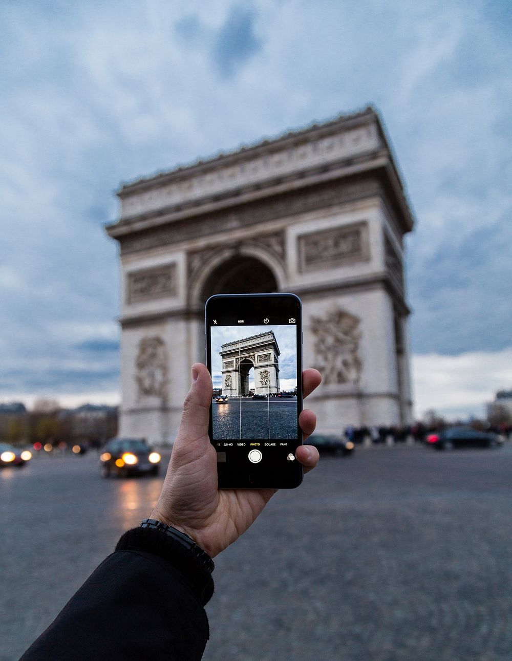 The hand of a man taking a photo of the Arc de Triomphe with his iPhone mobile smartphone while in traffic.. Original public…